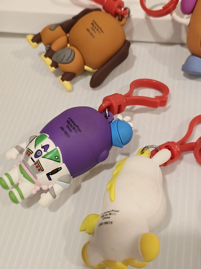 May include: A selection of character keyrings, featuring a brown dog, Buzz Lightyear, and a white unicorn. Each keyring has a red clasp. The keyrings are made from a rubbery material and are brightly coloured.