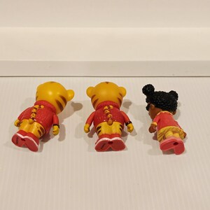 K&ouml;nnte beinhalten: Drei kleine Figuren von Zeichentrickfiguren. Zwei sind gelbe Tiger mit roten Jacken und gelben Hosen. Die dritte ist ein M&auml;dchen mit dunkler Haut und schwarzen Haaren, das ein rotes Kleid und rosa Schuhe tr&auml;gt. Alle stehen auf einer wei&szlig;en Oberfl&auml;che.