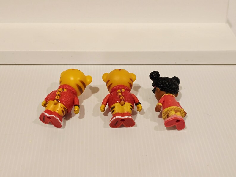 K&ouml;nnte beinhalten: Drei kleine Figuren von Zeichentrickfiguren. Zwei sind gelbe Tiger mit roten Jacken und gelben Hosen. Die dritte ist ein M&auml;dchen mit dunkler Haut und schwarzen Haaren, das ein rotes Kleid und rosa Schuhe tr&auml;gt. Alle stehen auf einer wei&szlig;en Oberfl&auml;che.
