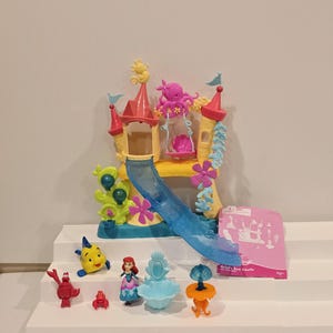 May include: Ariel's Sea Castle playset featuring a yellow, pink, and blue castle with a slide, swing, and various figurines. Includes Ariel, Flounder, Sebastian, and other characters. The playset is on a white surface.