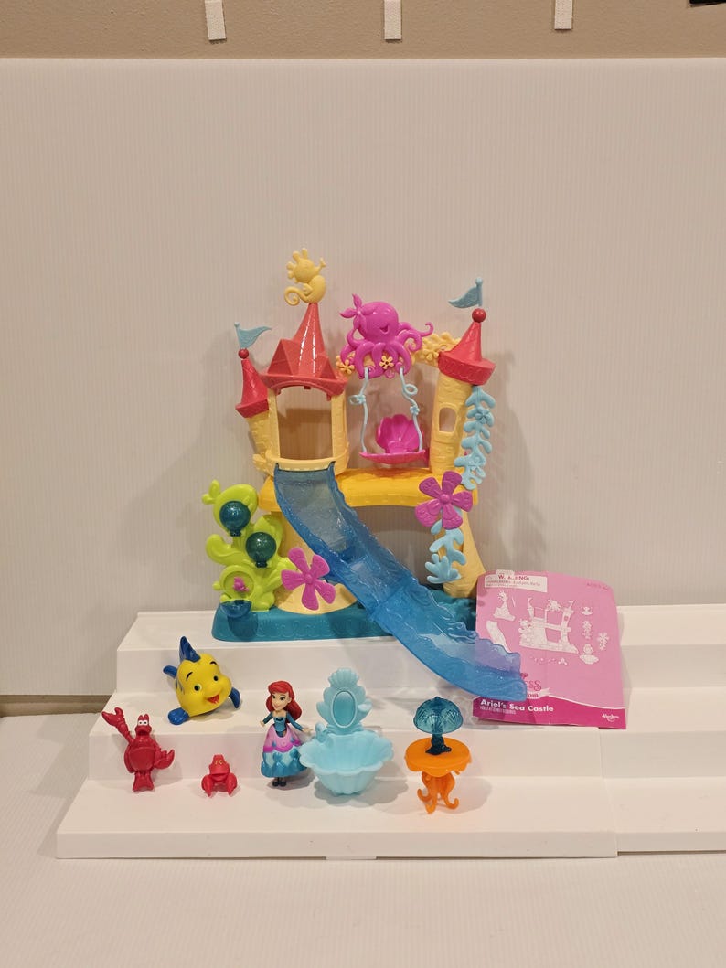 May include: Ariel's Sea Castle playset featuring a yellow, pink, and blue castle with a slide, swing, and various figurines. Includes Ariel, Flounder, Sebastian, and other characters. The playset is on a white surface.