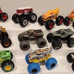 Op de afbeelding: Een verzameling speelgoed monstertrucks in verschillende kleuren, waaronder rood, geel, groen en bruin. Elke truck heeft grote, oversized banden en gedetailleerde ontwerpen. De trucks zijn op witte planken geplaatst.