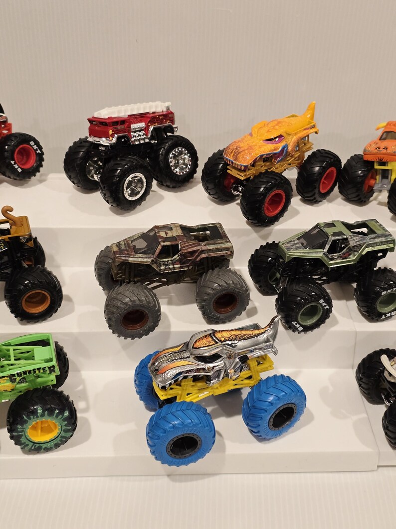 Op de afbeelding: Een verzameling speelgoed monstertrucks in verschillende kleuren, waaronder rood, geel, groen en bruin. Elke truck heeft grote, oversized banden en gedetailleerde ontwerpen. De trucks zijn op witte planken geplaatst.