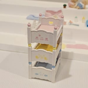 Puede incluir: Una cama litera en miniatura blanca con tres niveles. Cada nivel tiene un colch&oacute;n de diferente color: rosa, amarillo y azul. Los marcos de la cama est&aacute;n decorados con dise&ntilde;os florales y de animales. El nivel superior tiene un dise&ntilde;o de corona.