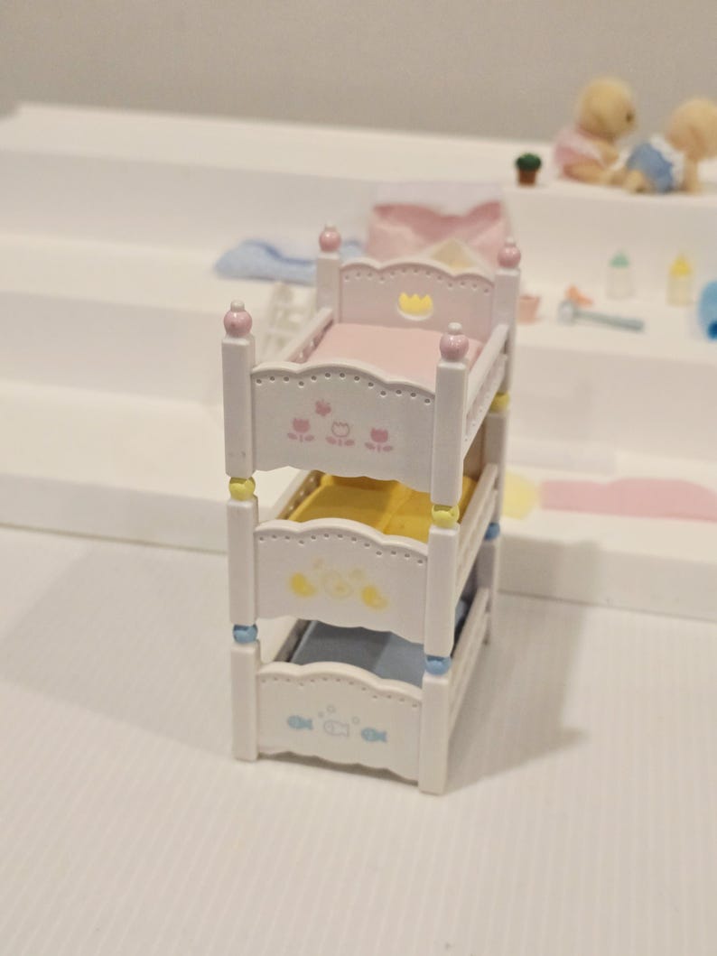 Puede incluir: Una cama litera en miniatura blanca con tres niveles. Cada nivel tiene un colch&oacute;n de diferente color: rosa, amarillo y azul. Los marcos de la cama est&aacute;n decorados con dise&ntilde;os florales y de animales. El nivel superior tiene un dise&ntilde;o de corona.