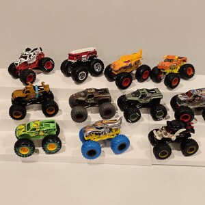 Op de afbeelding: Een verzameling speelgoed monstertrucks in verschillende kleuren en ontwerpen. De trucks worden gepresenteerd op een wit, trapsgewijs platform. Elke truck heeft grote, oversized wielen en gedetailleerde graphics.