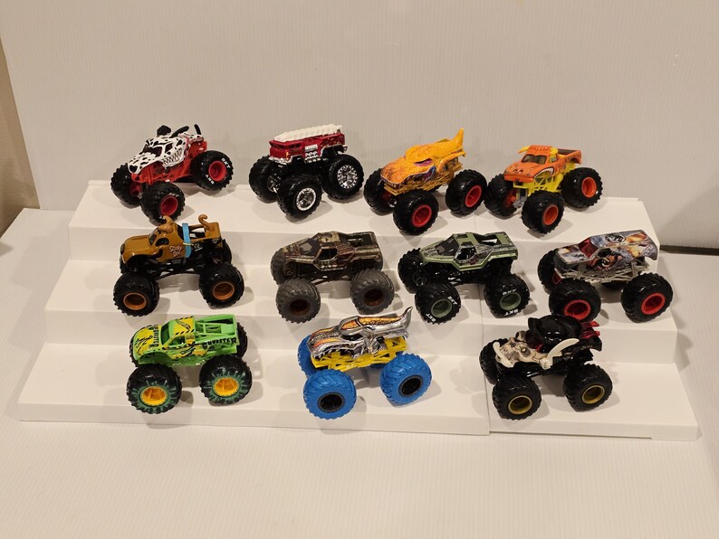 Op de afbeelding: Een verzameling speelgoed monstertrucks in verschillende kleuren en ontwerpen. De trucks worden gepresenteerd op een wit, trapsgewijs platform. Elke truck heeft grote, oversized wielen en gedetailleerde graphics.