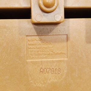 May include: Close-up of a tan plastic object with a textured surface. The text "EPOCH MADE IN CHINA" is visible, along with the manufacturer's address in Tokyo, Japan. The serial number A07618 is also printed on the surface.