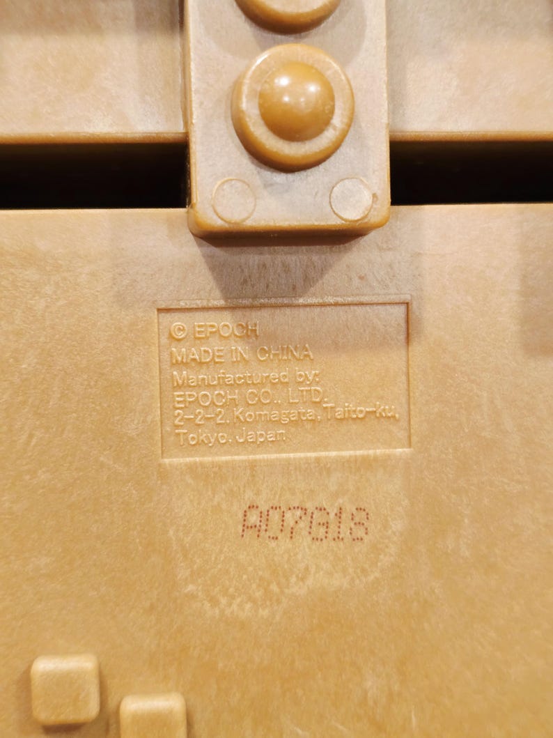 May include: Close-up of a tan plastic object with a textured surface. The text "EPOCH MADE IN CHINA" is visible, along with the manufacturer's address in Tokyo, Japan. The serial number A07618 is also printed on the surface.