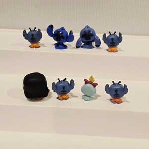 May include: A collection of small, colorful figurines featuring the character Stitch from the animated film Lilo & Stitch. The figurines are primarily blue with orange accents, and one is light blue with a red bow.