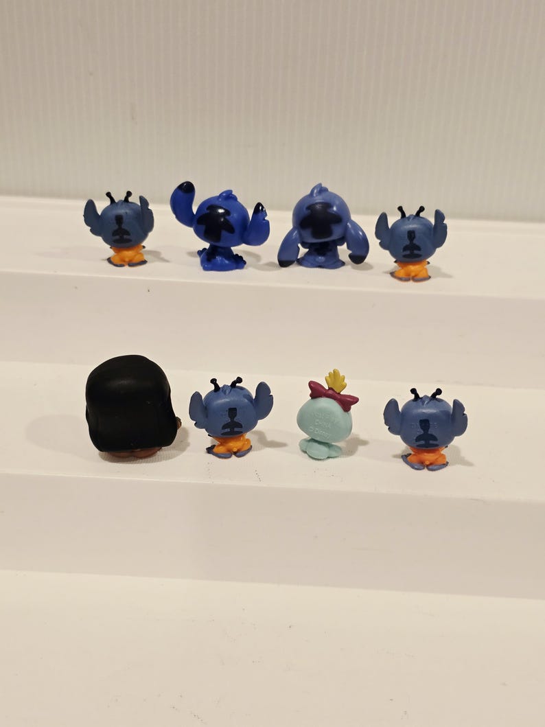 May include: A collection of small, colorful figurines featuring the character Stitch from the animated film Lilo & Stitch. The figurines are primarily blue with orange accents, and one is light blue with a red bow.
