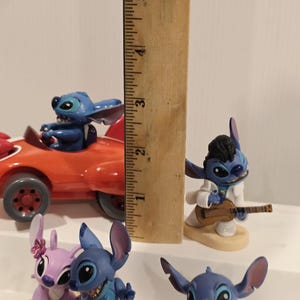 May include: A collection of Stitch figurines, including one in a red car, one playing a guitar, and others in various poses. The tallest figurine measures approximately 5 inches.