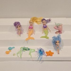 May include: A collection of miniature mermaid and fairy dolls with accessories. The dolls have colorful hair and outfits, with mermaid tails and fairy wings. Accessories include a starfish, a dolphin figurine, and hair pieces.