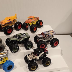 Op de afbeelding: Een verzameling speelgoed monstertrucks in verschillende kleuren, waaronder oranje, geel, groen en zwart. Elke truck heeft grote wielen met rode of gouden accenten. De trucks worden getoond op een witte, getrapte ondergrond.