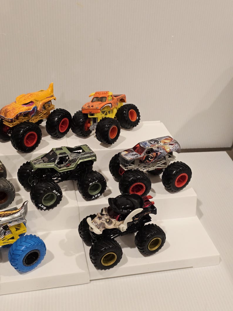 Op de afbeelding: Een verzameling speelgoed monstertrucks in verschillende kleuren, waaronder oranje, geel, groen en zwart. Elke truck heeft grote wielen met rode of gouden accenten. De trucks worden getoond op een witte, getrapte ondergrond.