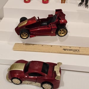 May include: Two toy cars, one a red race car with gold wheels and accents, and the other a red sports car with gold accents. A wooden ruler with the text "-WESTCOTT Since 1872-" is also present.
