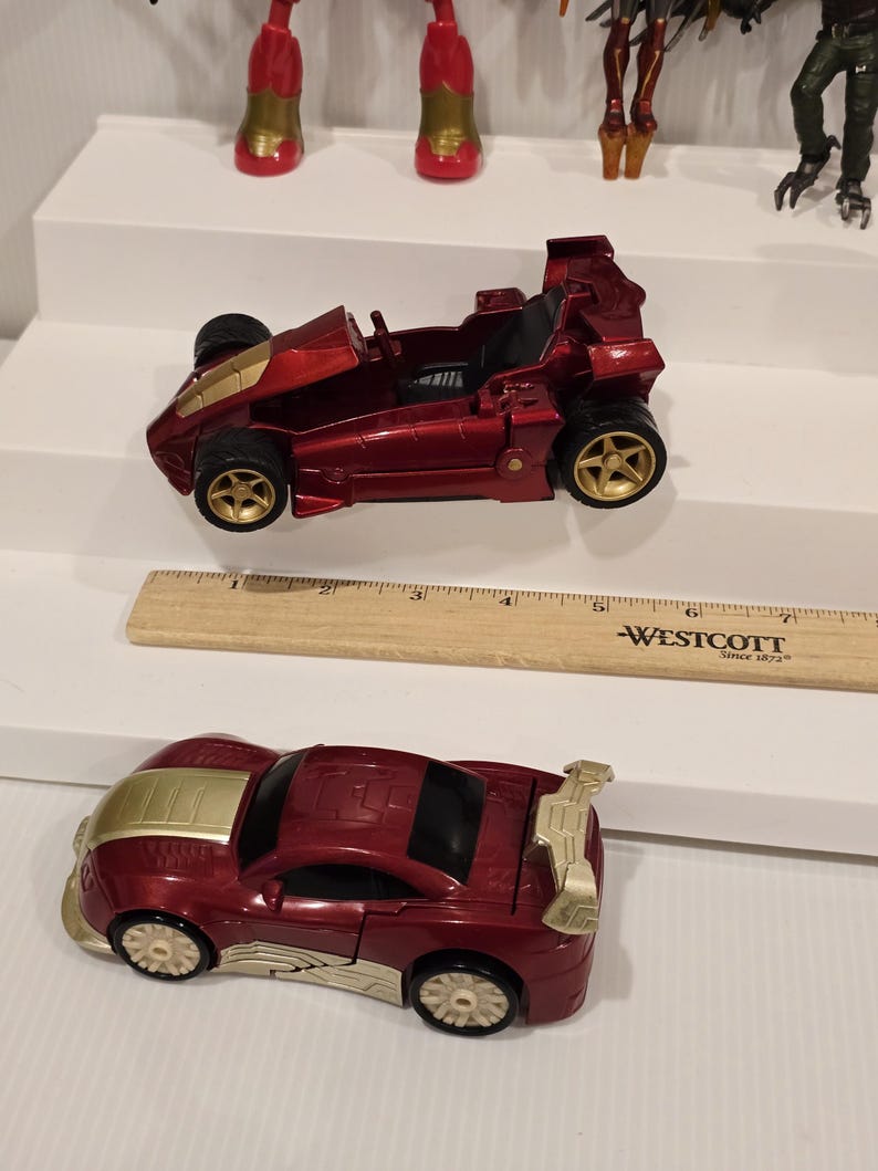 May include: Two toy cars, one a red race car with gold wheels and accents, and the other a red sports car with gold accents. A wooden ruler with the text "-WESTCOTT Since 1872-" is also present.