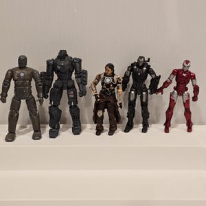 May include: A collection of six action figures of various characters, including Iron Man, War Machine, and other armored figures. The figures are in various colors, including red, black, and silver. The figures are standing on a white surface.