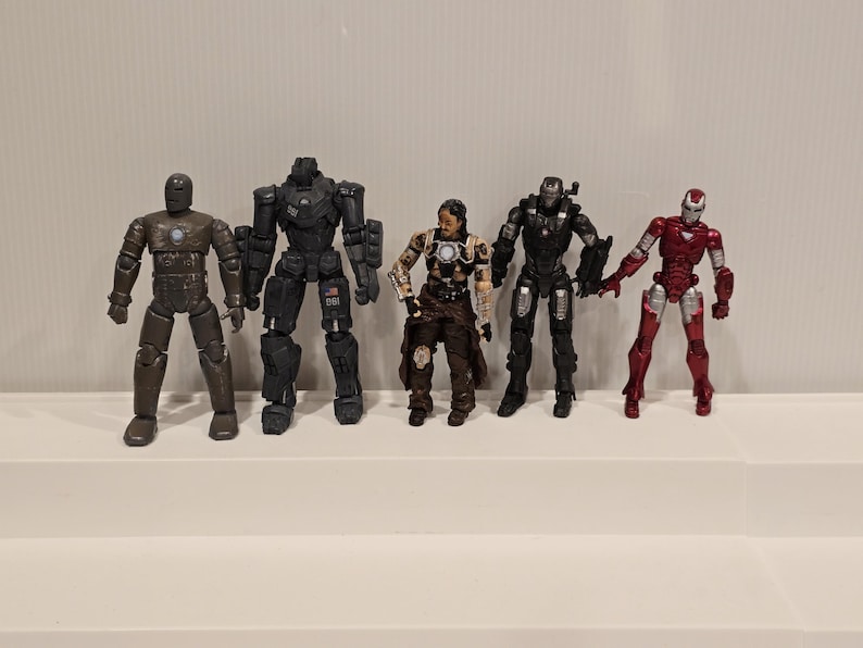 May include: A collection of six action figures of various characters, including Iron Man, War Machine, and other armored figures. The figures are in various colors, including red, black, and silver. The figures are standing on a white surface.