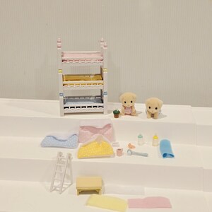 Puede incluir: Un juego de dormitorio de casa de mu&ntilde;ecas en miniatura con una litera, dos cachorros de juguete y varios accesorios. La litera tiene ropa de cama rosa, amarilla y azul. Los accesorios incluyen una escalera, una mesa, mantas, biberones y un martillo de juguete.