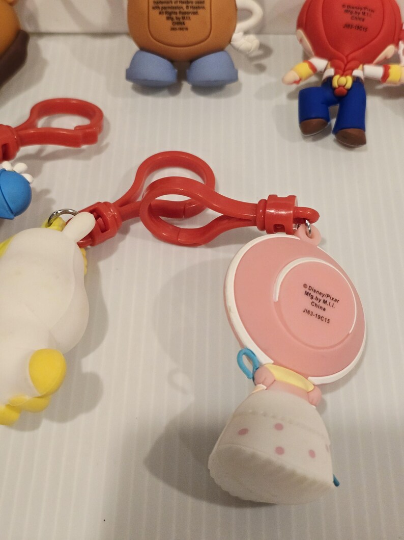 May include: A selection of Toy Story character keyrings. The keyrings feature characters such as Bo Peep, Mr. Potato Head, and Woody. The keyrings are made of plastic and have red plastic clips.