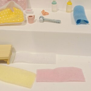 Puede incluir: Una colecci&oacute;n de art&iacute;culos de cuidado para beb&eacute;s en miniatura, que incluyen biberones, un cepillo de dientes, un pa&ntilde;o y un chupete. Tambi&eacute;n se ven un cambiador amarillo con lunares y un pa&ntilde;al rosa y blanco. Los art&iacute;culos est&aacute;n dispuestos sobre una superficie blanca.
