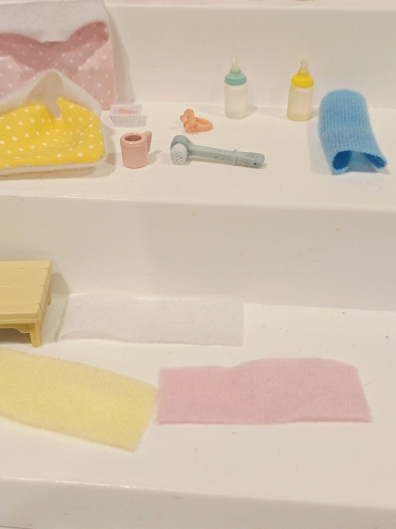 Puede incluir: Una colecci&oacute;n de art&iacute;culos de cuidado para beb&eacute;s en miniatura, que incluyen biberones, un cepillo de dientes, un pa&ntilde;o y un chupete. Tambi&eacute;n se ven un cambiador amarillo con lunares y un pa&ntilde;al rosa y blanco. Los art&iacute;culos est&aacute;n dispuestos sobre una superficie blanca.