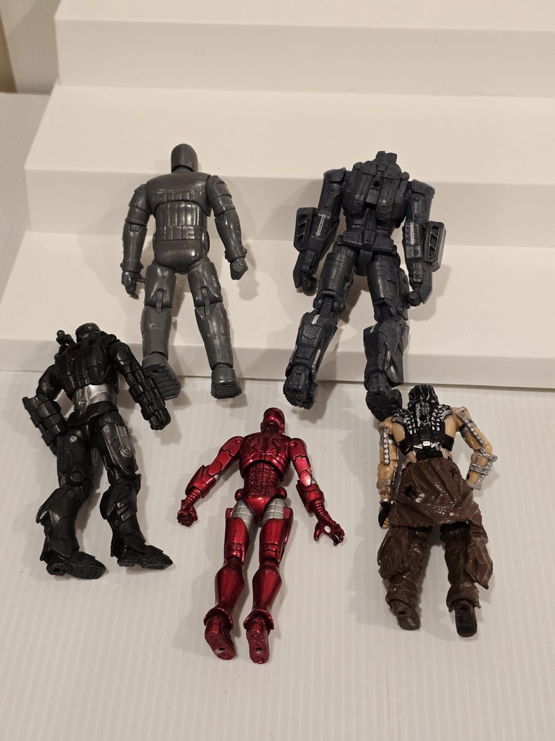 May include: Six action figures are arranged on a white surface. The figures include a metallic gray robot, a red and silver figure, and several black figures with varying armor designs. The figures are detailed and appear to be from a science fiction or superhero theme.