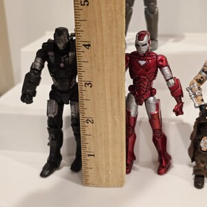 May include: Two action figures of superheroes, one black and silver, the other red and silver, are displayed next to a wooden ruler. The figures are approximately 3 inches tall.