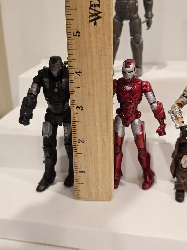 May include: Two action figures of superheroes, one black and silver, the other red and silver, are displayed next to a wooden ruler. The figures are approximately 3 inches tall.