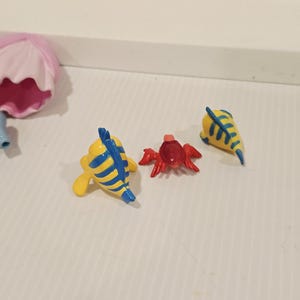 K&ouml;nnte beinhalten: Eine Sammlung kleiner, farbenfroher Figuren. Das Set enth&auml;lt zwei gelbe Fische mit blauen Streifen, eine rote Krabbe und eine gelbe fischf&ouml;rmige Figur mit blauen Streifen. Die Figuren sind wahrscheinlich Spielzeug oder Sammlerst&uuml;cke.
