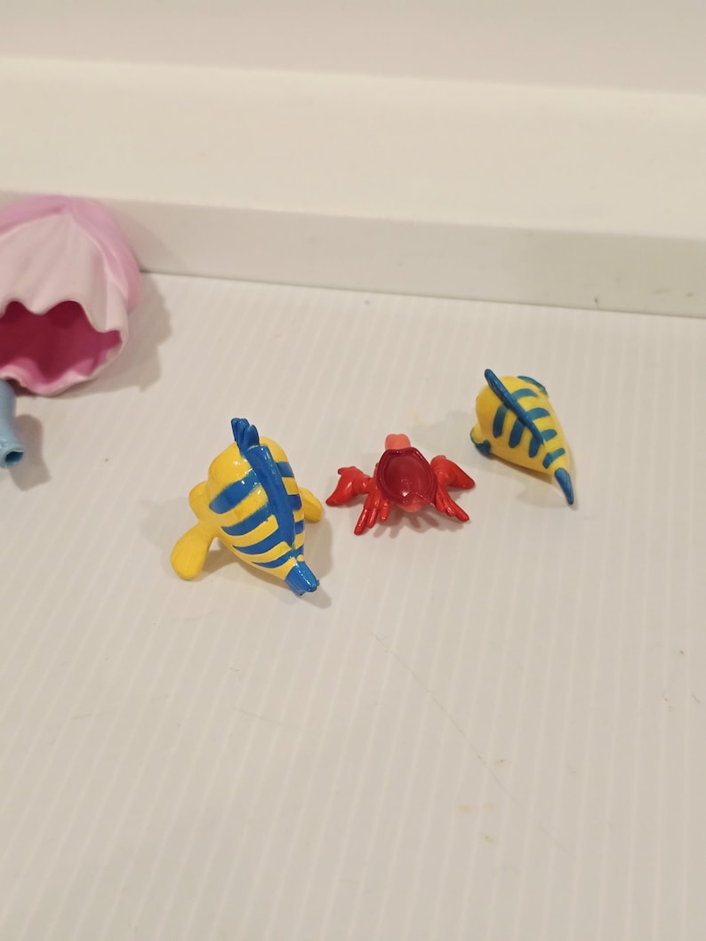 K&ouml;nnte beinhalten: Eine Sammlung kleiner, farbenfroher Figuren. Das Set enth&auml;lt zwei gelbe Fische mit blauen Streifen, eine rote Krabbe und eine gelbe fischf&ouml;rmige Figur mit blauen Streifen. Die Figuren sind wahrscheinlich Spielzeug oder Sammlerst&uuml;cke.