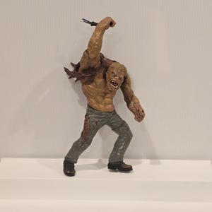 May include: A detailed figurine of a monstrous creature with exposed muscles and a menacing expression. The figure is primarily brown with grey pants and black boots. It is holding a small, dark object in its raised right hand.