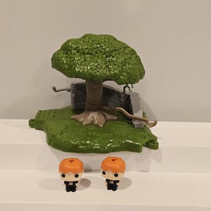 May include: A green plastic playset featuring a tree, stone structure, and two figurines with orange hair and black outfits. The tree has a full, textured green canopy. The figurines are in a stylized, collectible design.