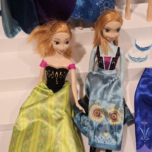 May include: Two Anna dolls from the film Frozen. One doll is dressed in a green and yellow dress with pink floral details and a black bodice. The other doll wears a blue dress with sunflower designs and black boots. Other doll parts are visible in the background.