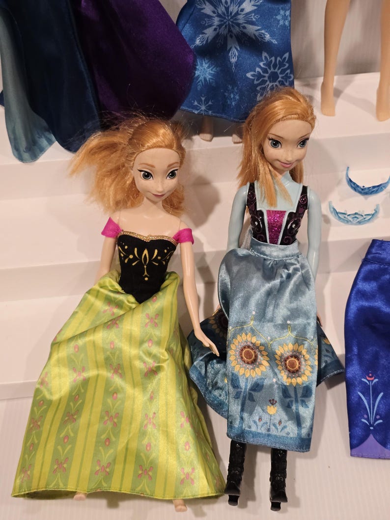 May include: Two Anna dolls from the film Frozen. One doll is dressed in a green and yellow dress with pink floral details and a black bodice. The other doll wears a blue dress with sunflower designs and black boots. Other doll parts are visible in the background.
