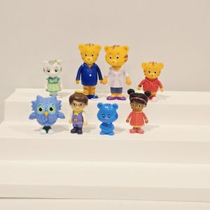 May include: A collection of eight small plastic character figurines including a blue owl, a cat in a green dress, two tiger figures in blue and white tops, a tiger in a red hoodie, a human figure with glasses, a blue bear, and a girl in a red dress.