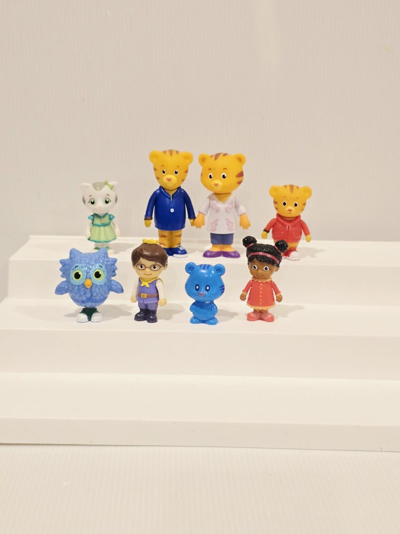 May include: A collection of eight small plastic character figurines including a blue owl, a cat in a green dress, two tiger figures in blue and white tops, a tiger in a red hoodie, a human figure with glasses, a blue bear, and a girl in a red dress.