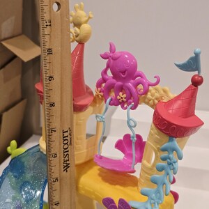 May include: A colorful toy castle with a ruler for scale. The castle features pink, yellow, and blue accents, including a pink octopus on a swing, a yellow sea creature, and a blue flag. The ruler shows the castle is approximately 12 inches tall.