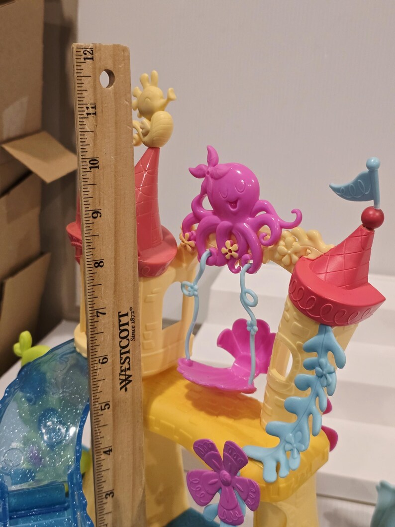 May include: A colorful toy castle with a ruler for scale. The castle features pink, yellow, and blue accents, including a pink octopus on a swing, a yellow sea creature, and a blue flag. The ruler shows the castle is approximately 12 inches tall.