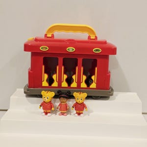 K&ouml;nnte beinhalten: Ein roter Spielzeugwagen mit gelbem Griff und drei transparenten Fenstern. Vor dem Wagen stehen drei kleine Figuren von Zeichentrickfiguren. Die Figuren tragen rote Jacken und Kleider.