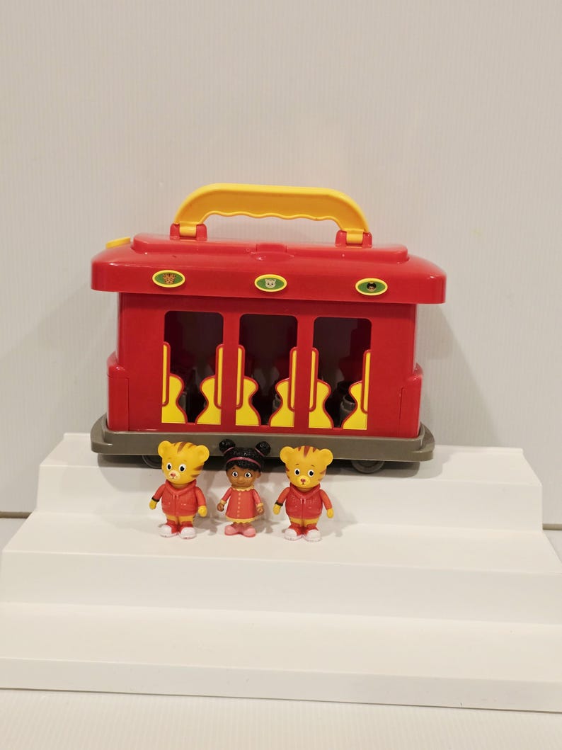 K&ouml;nnte beinhalten: Ein roter Spielzeugwagen mit gelbem Griff und drei transparenten Fenstern. Vor dem Wagen stehen drei kleine Figuren von Zeichentrickfiguren. Die Figuren tragen rote Jacken und Kleider.