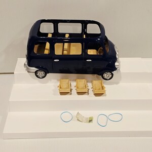 May include: A dark blue toy bus with a light tan interior and three matching chairs. The bus has silver-colored wheels and trim. Two blue rubber bands and a small paper are also present.