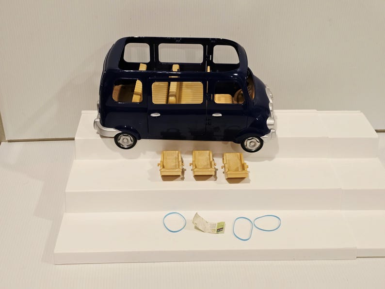 May include: A dark blue toy bus with a light tan interior and three matching chairs. The bus has silver-colored wheels and trim. Two blue rubber bands and a small paper are also present.