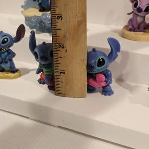 May include: A collection of blue Stitch figurines, some holding hearts, others in various poses. A wooden ruler shows the height of one figurine is approximately 2 inches. The figurines are on a white surface.