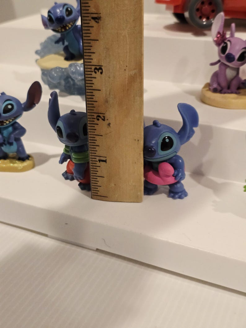 May include: A collection of blue Stitch figurines, some holding hearts, others in various poses. A wooden ruler shows the height of one figurine is approximately 2 inches. The figurines are on a white surface.