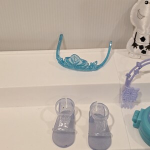 K&ouml;nnte beinhalten: Ein hellblaues Kunststoff-Diadem mit Schneeflockenmuster und zwei Paar durchsichtige Kunststoff-Sandalen. Die Sandalen haben einen Kn&ouml;chelriemen und einen Zehenriemen. Die Artikel sind Teil eines Kinderspielzeugsets.