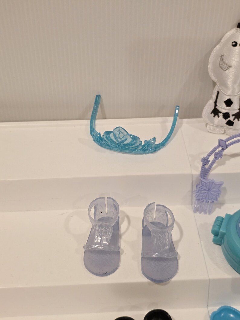 K&ouml;nnte beinhalten: Ein hellblaues Kunststoff-Diadem mit Schneeflockenmuster und zwei Paar durchsichtige Kunststoff-Sandalen. Die Sandalen haben einen Kn&ouml;chelriemen und einen Zehenriemen. Die Artikel sind Teil eines Kinderspielzeugsets.