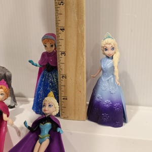 May include: A collection of small plastic figurines of characters from the animated film "Frozen". The figures include Elsa, Anna, and other characters. A wooden ruler is placed next to the figurines to show their height, approximately 7.6 cm.