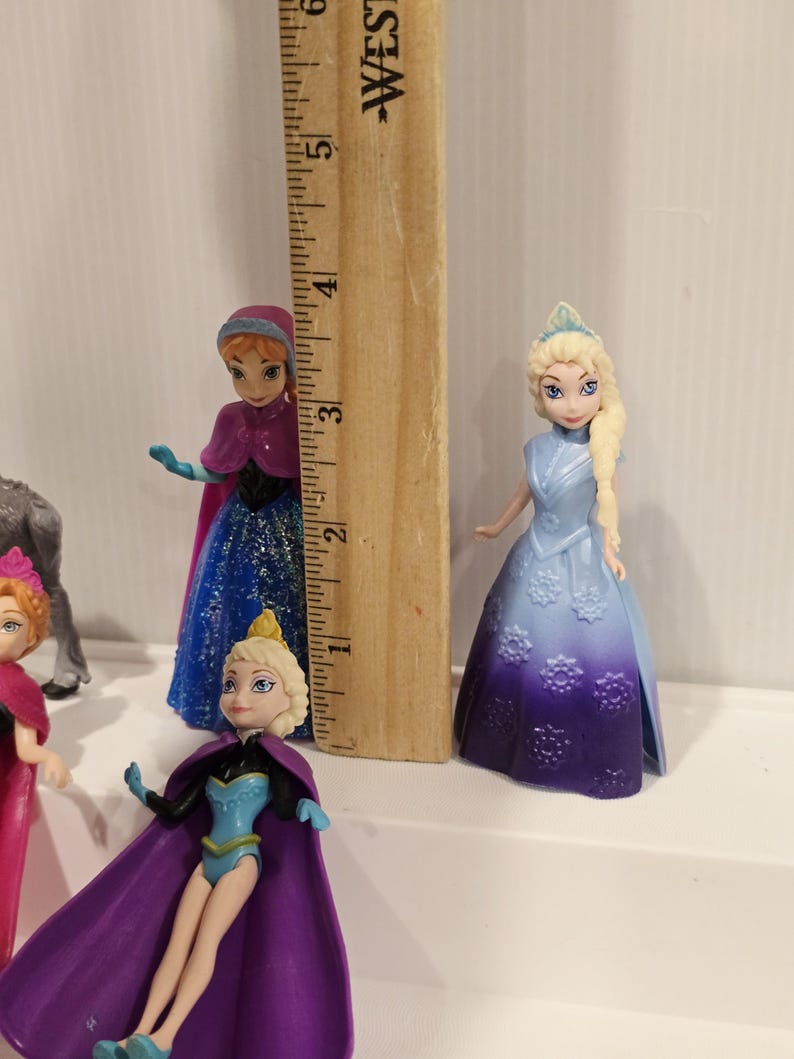 May include: A collection of small plastic figurines of characters from the animated film "Frozen". The figures include Elsa, Anna, and other characters. A wooden ruler is placed next to the figurines to show their height, approximately 7.6 cm.